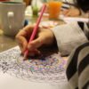The hand of a person coloring a mandala design using a pink colored pencil