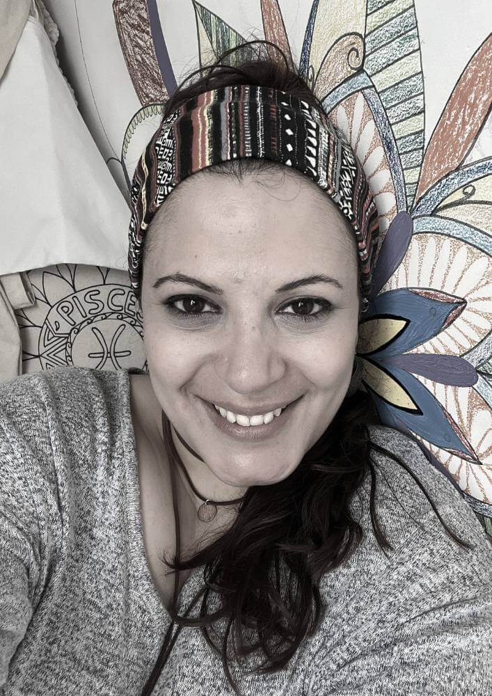 this is the image of a smiling woman wearing a boho head bandana, and has a mural mandala background and the drawing of Pisces behind her